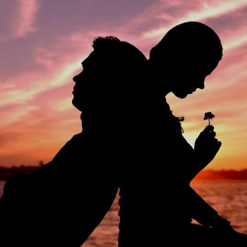 A silhouette of a couple sitting back to back thinking of how to say I love you in Spanish with a beautiful sunset in the background